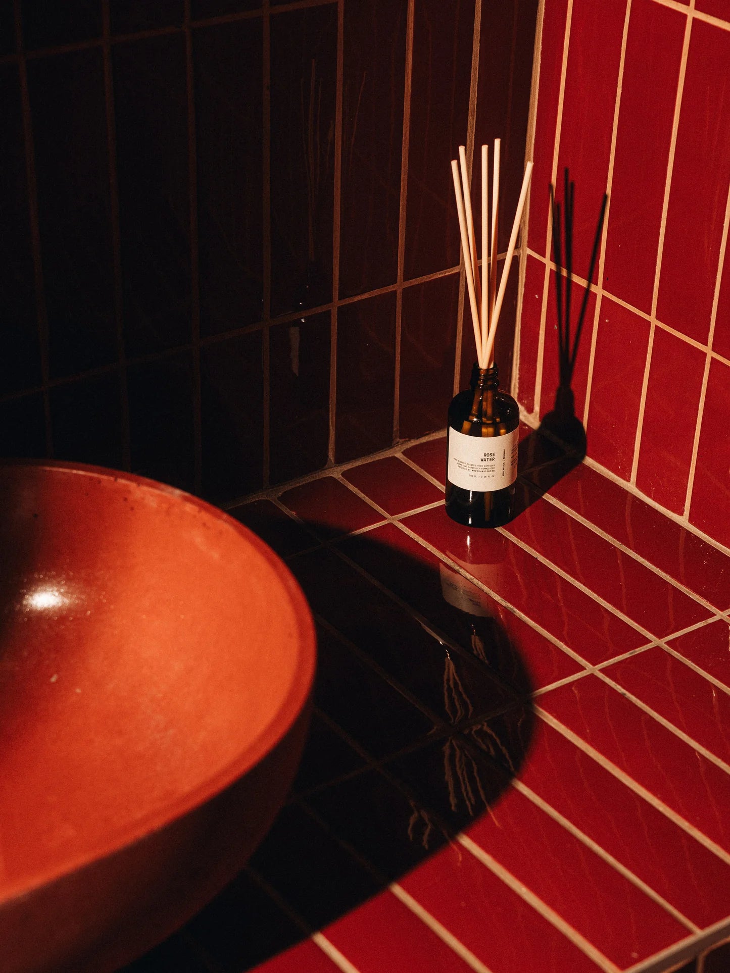 Bottle of diffuser with reeds on a red tiled wall next to a ceramic bowl.