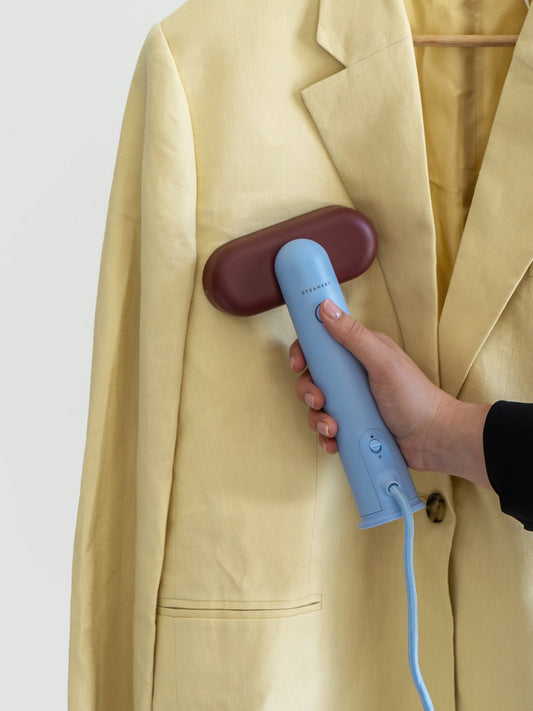 Hand using a blue handheld steamer on a beige coat against a white background