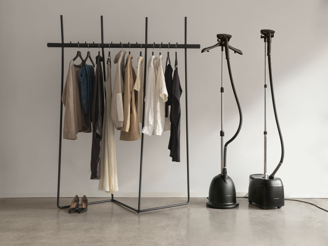 Clothing rack with hanging clothes next to two steaming irons on a plain background