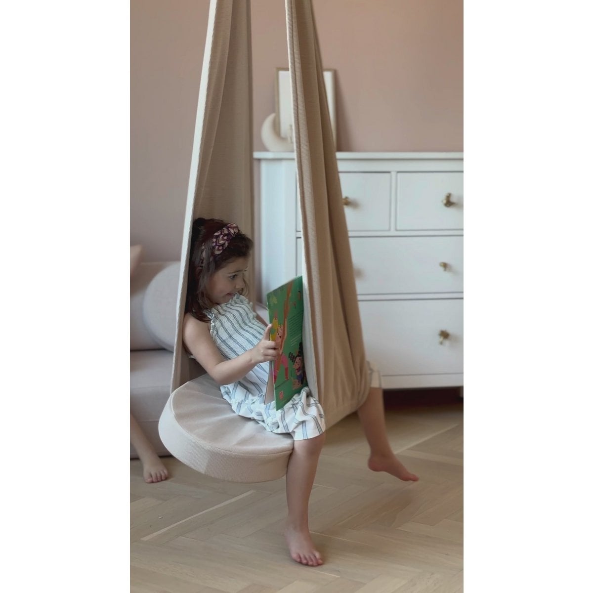 Child sitting in a hanging chair reading a book in a room with a dresser.