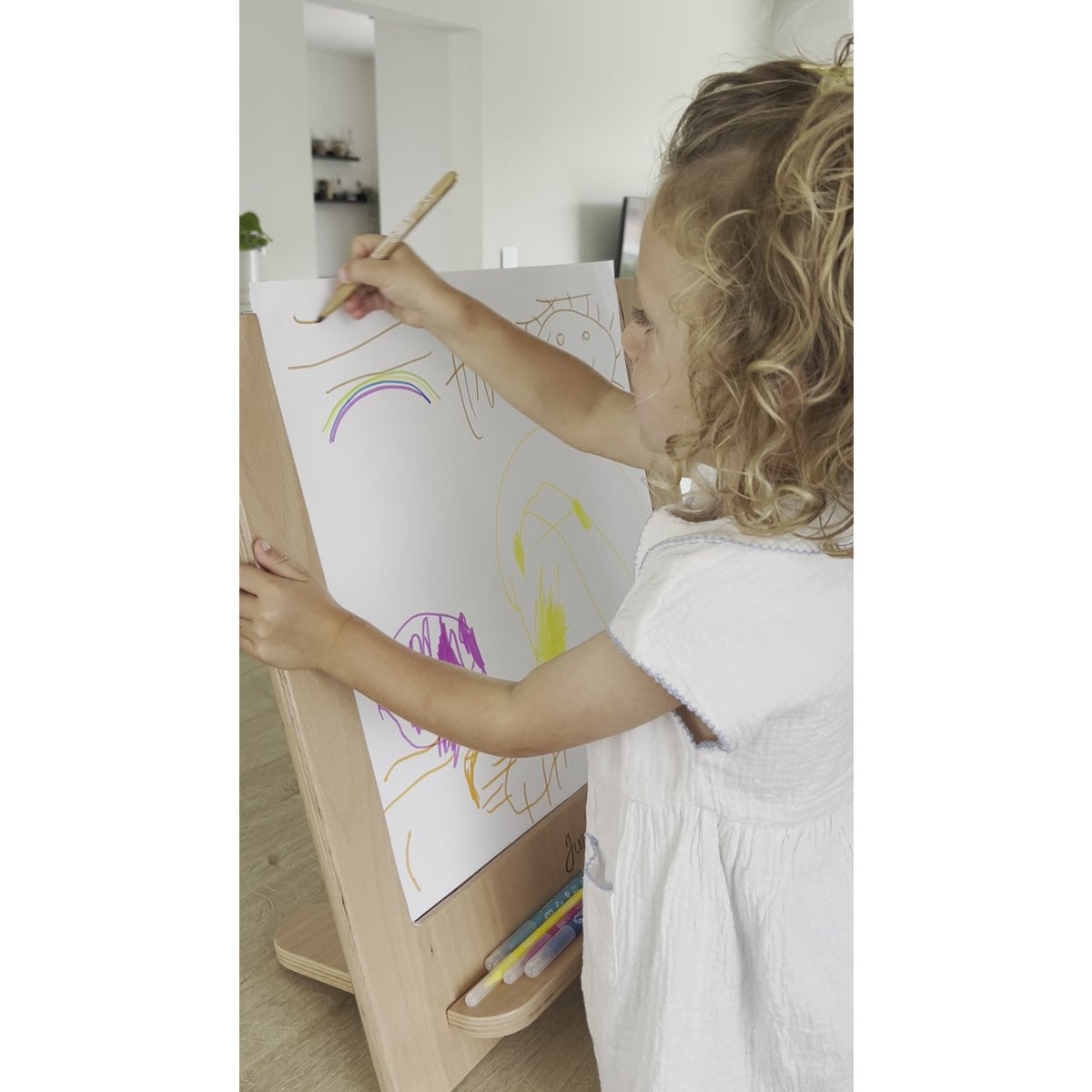 Child drawing on a white board with colorful crayons in a minimalistic room.