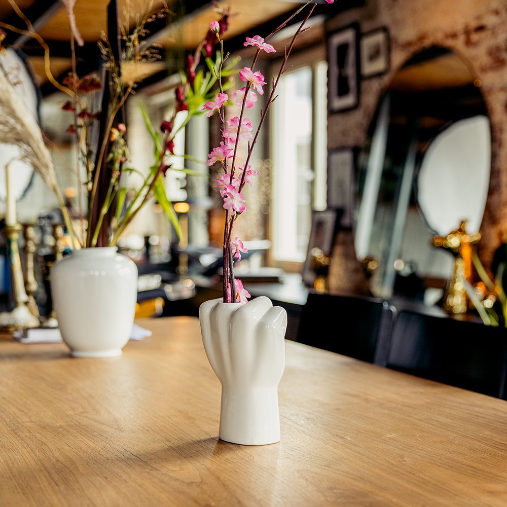 White hand-shaped vase with pink flowers on a wooden table in a blurred indoor setting