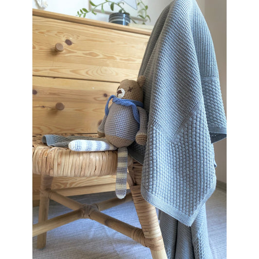 Knitted teddy bear and blanket on a wicker chair with wooden dresser in the background