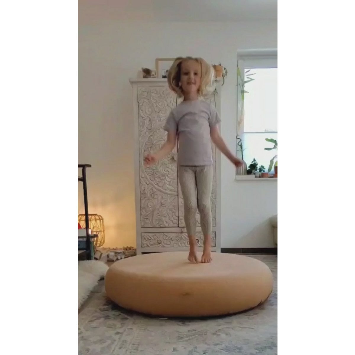 Child standing on a round beige cushion in a room with decorative elements.