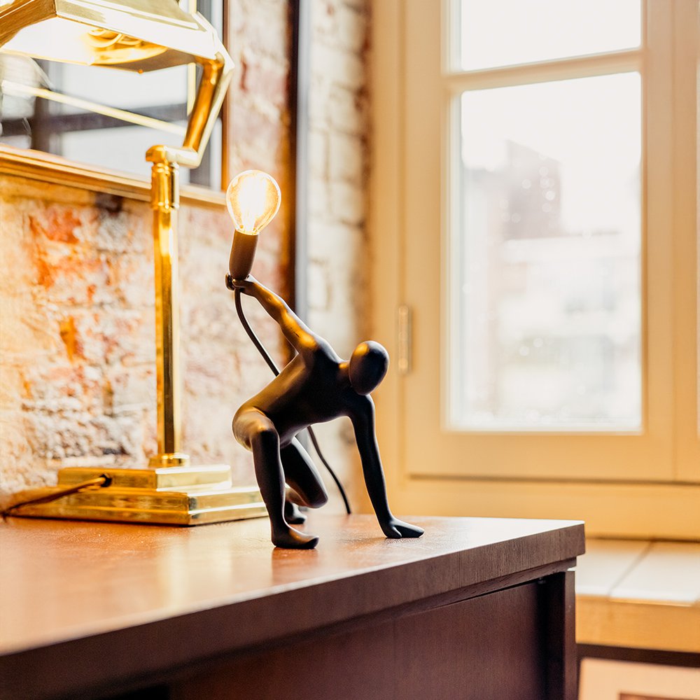 Decorative sculpture of a person holding a light bulb on a wooden surface with a lamp in the background.