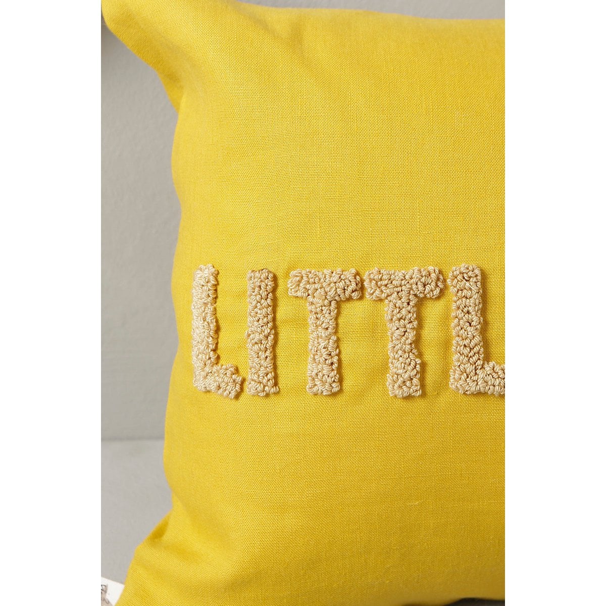 Yellow pillow with 'LITTLE' embroidered in beige letters on a neutral background