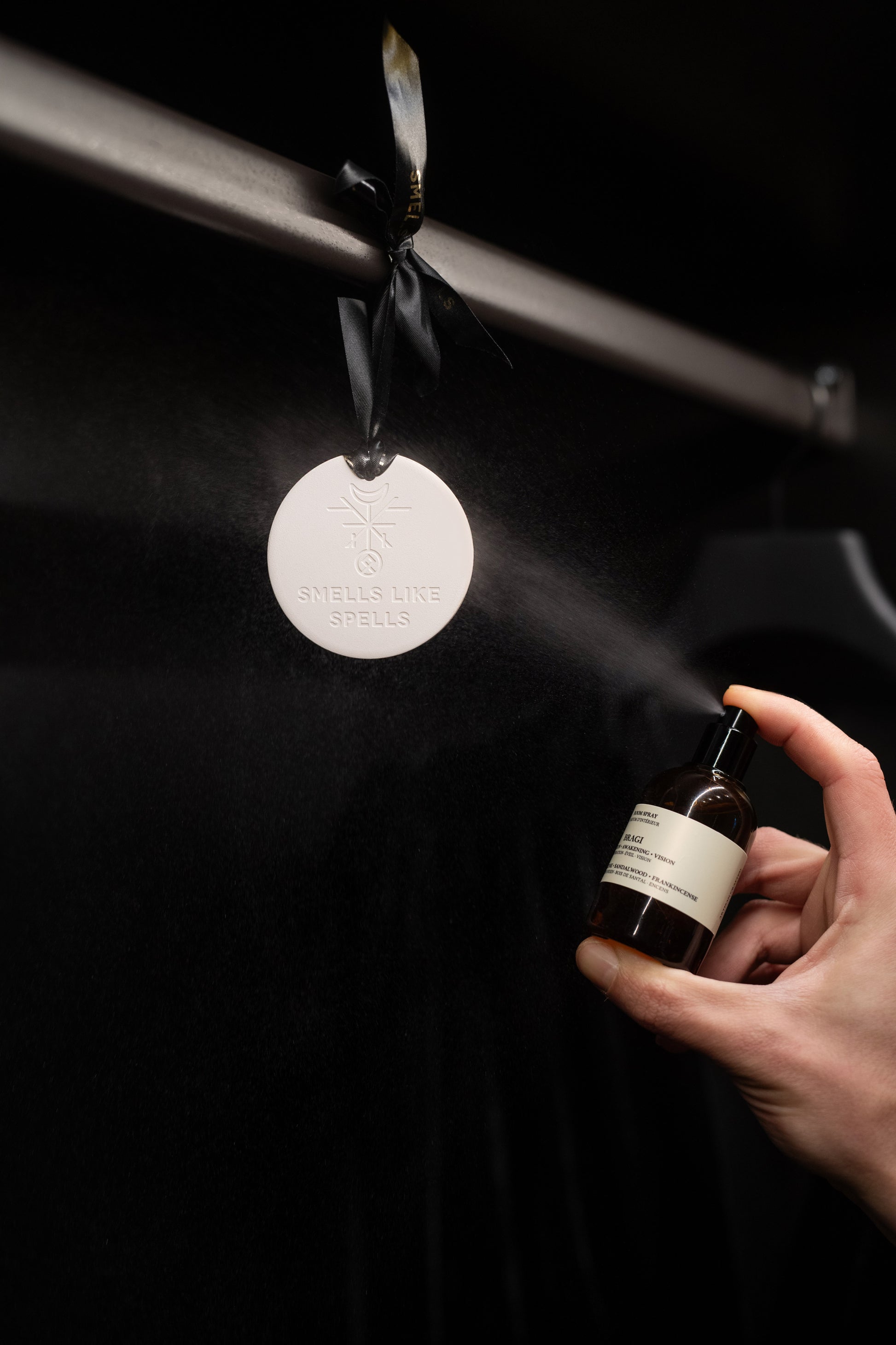 Hand holding a bottle of perfume next to a hanging metal disc with branding on a dark background