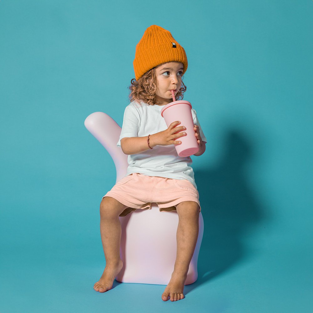 Child sitting on a pink chair, drinking from a pink cup against a blue background