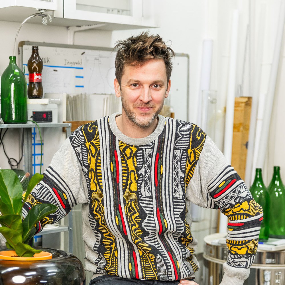 Klaas Kuiken wearing a patterned sweater in a kitchen setting with bottles and plants.
