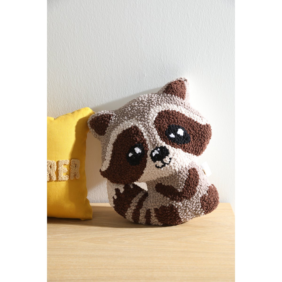 Plush raccoon toy on a wooden surface with a yellow pillow in the background