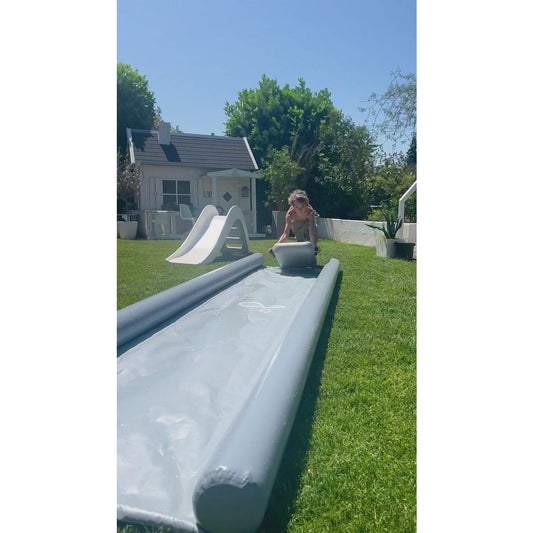 Child sliding down a large inflatable slide in a backyard with a small playhouse and trees in the background.