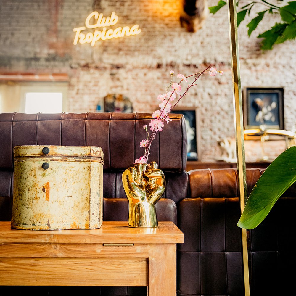 Decorative setting with a brown leather sofa, wooden table, and decorative items in a room with 'Club Tropicana' neon sign.