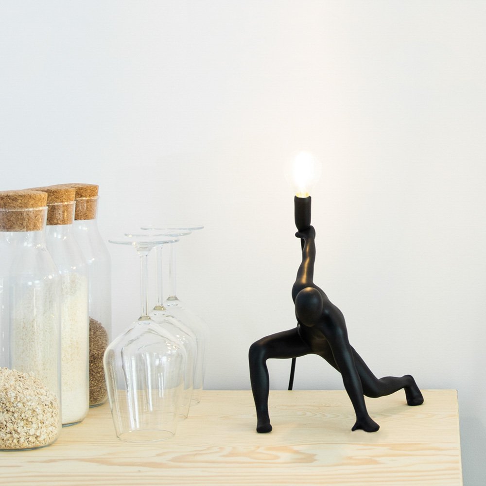 Black candle holder shaped like a person in a yoga pose on a wooden surface with jars and glasses in the background.