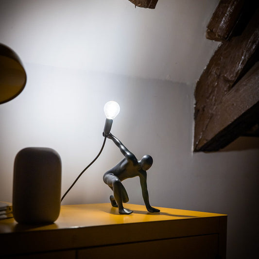 Decorative lamp shaped like a person holding a light bulb on a wooden surface.