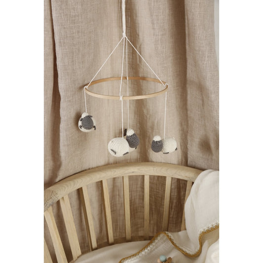 Baby crib with a hanging mobile featuring three knitted sheep against a beige curtain background.