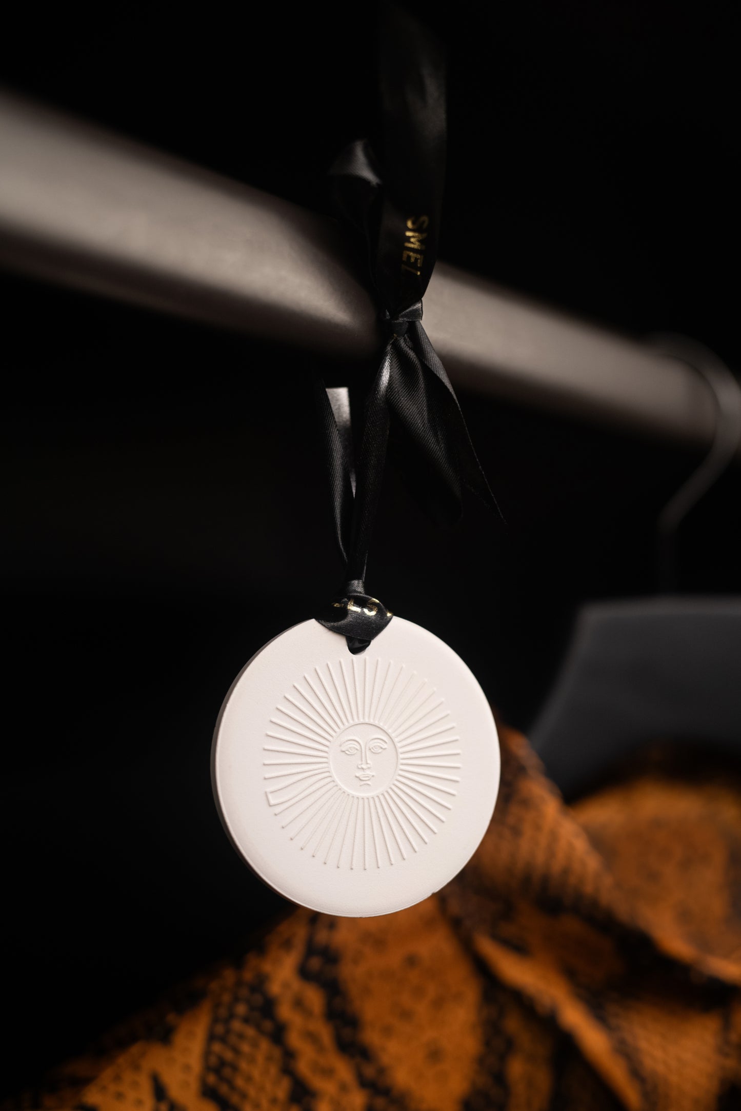 White decorative ornament with sun design hanging on a black ribbon against a dark background.