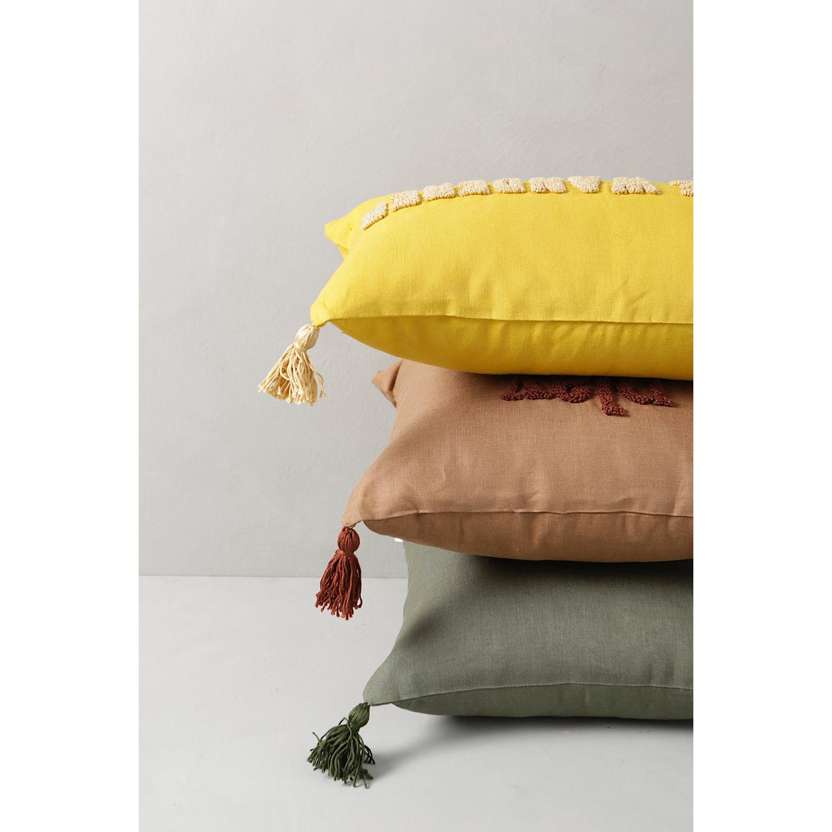 Stack of three decorative pillows in yellow, brown, and green with tassels on a light gray background.