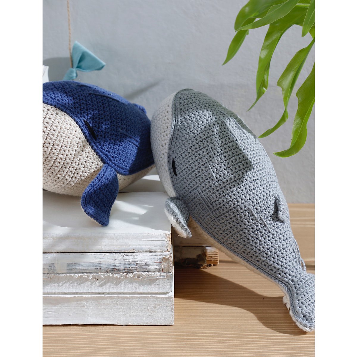 Two crocheted fish toys, one blue and one gray, on a wooden surface with a plant in the background.