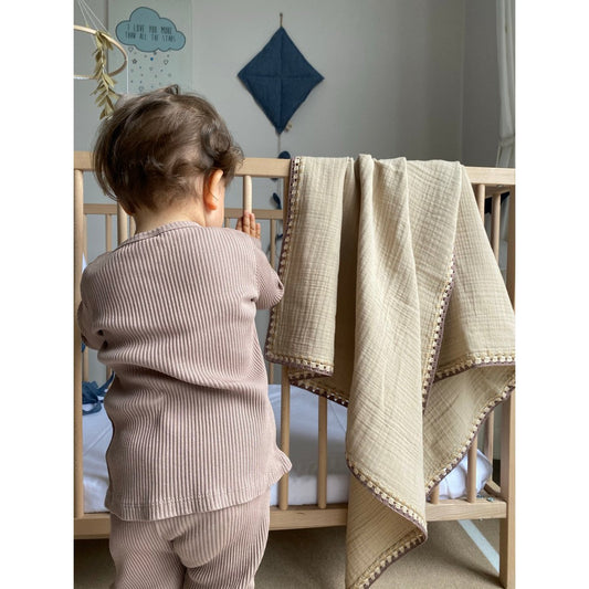 Child standing next to a crib with a beige blanket draped over the side in a nursery setting.