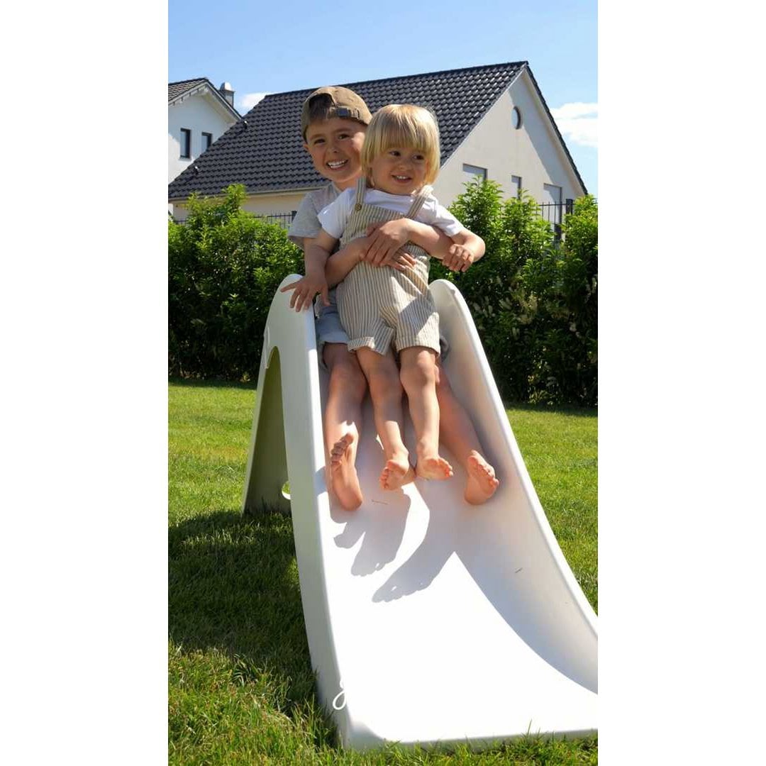 Two children sliding down a white slide in a backyard with a house and trees in the background.