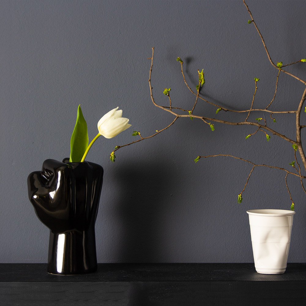 Black vase shaped like a hand holding a white tulip against a dark gray wall.
