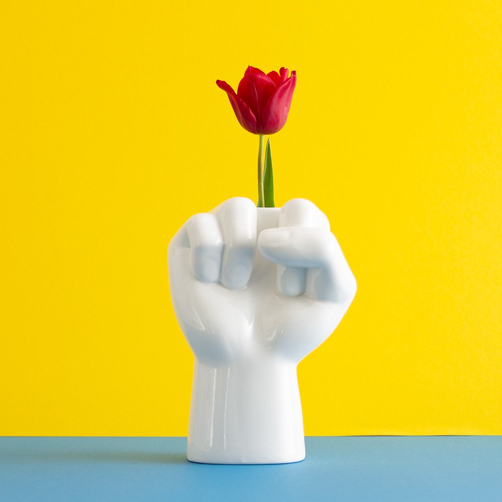 White ceramic vase shaped like a fist holding a red tulip against a yellow background