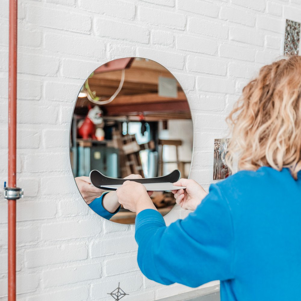 Job van den Berg a blue sweater using a hair straightener in front of a round mirror on a white brick wall.