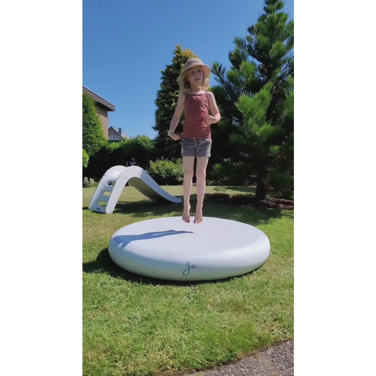 Child standing on a large inflatable disc in a backyard with a slide and trees in the background.